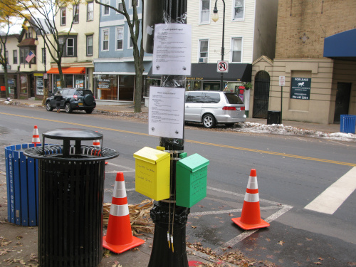 Photo of a light pole with a green mail box and a yellow mail box affixed to it. Two laminated paper signs are taped above them.