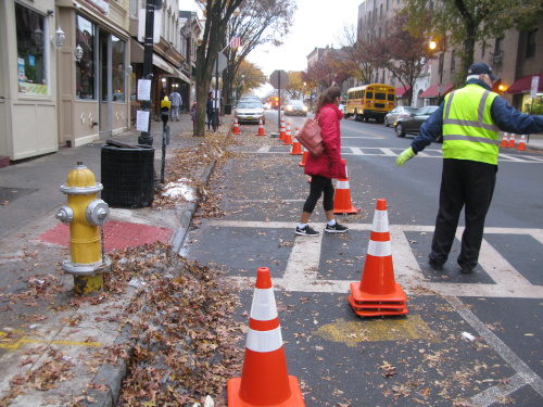 Photo of a T intersection of downtown street. Cones block off several parking spaces. The back of a stop sign on a temporary pole is at the far side of the coned area. A woman is walking across the street and a crossing guard holds up their arm.